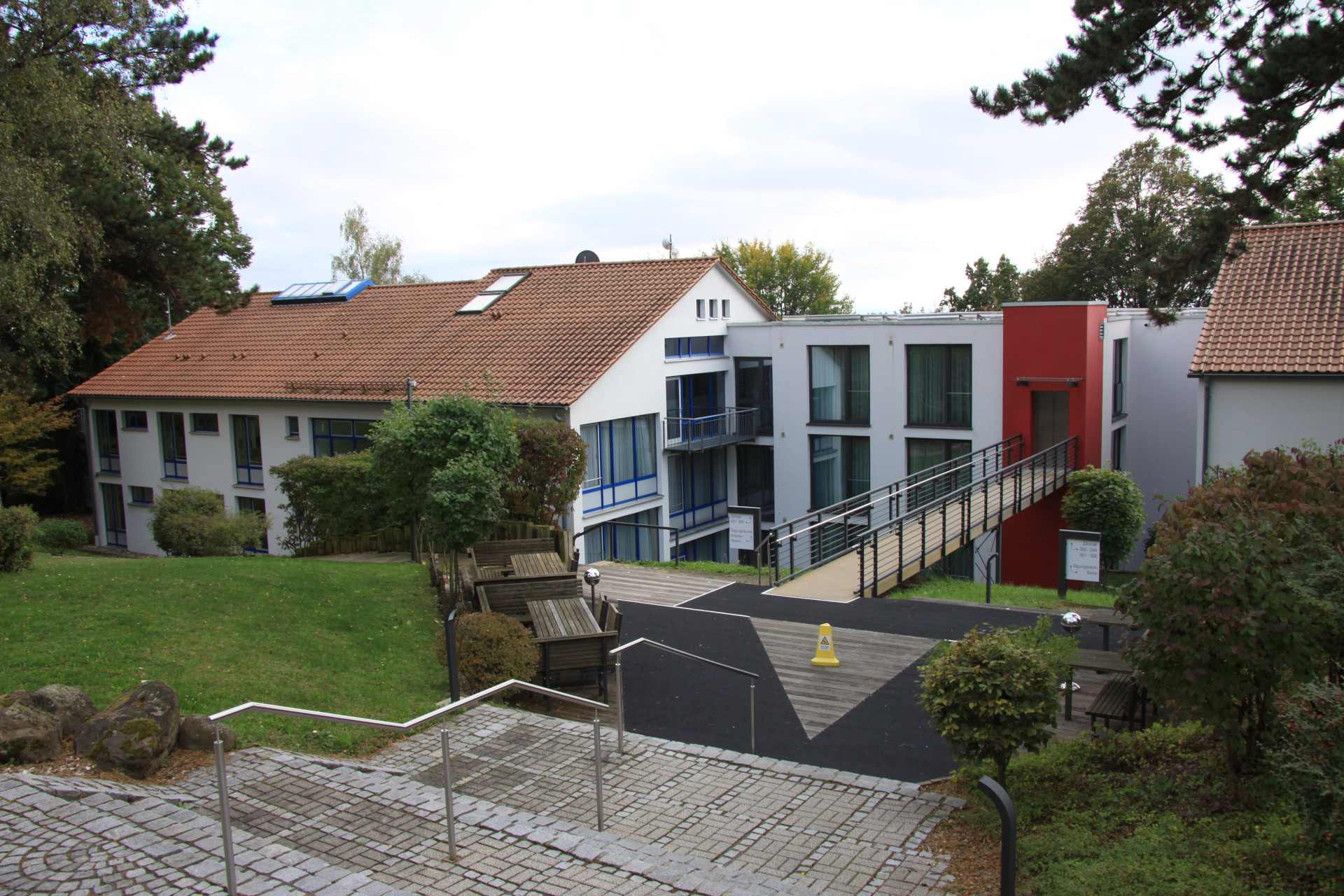 This picture shows two of the buildings with the rooms for visitors of the Seminarzentrum RÜckersbach. A smaller on connects both with an elevator and a bridge to the front there some stairs lead to the photograph. The buildings are surrounded by grass and trees. The sky is cloudy.