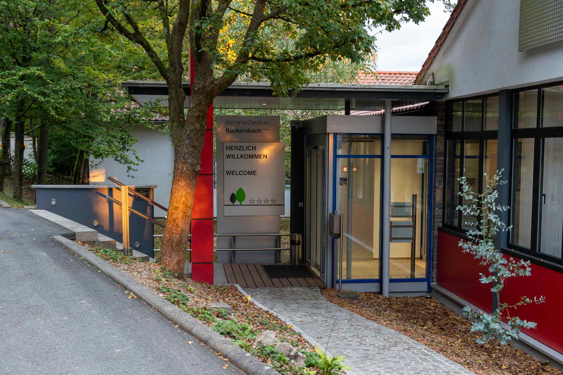 The picture shows the main entrance to the Seminarzentrum Rückersbach, the event location of osco. Warm sunlight shines through some trees behind the photograph. A sign welcomes the visitors.