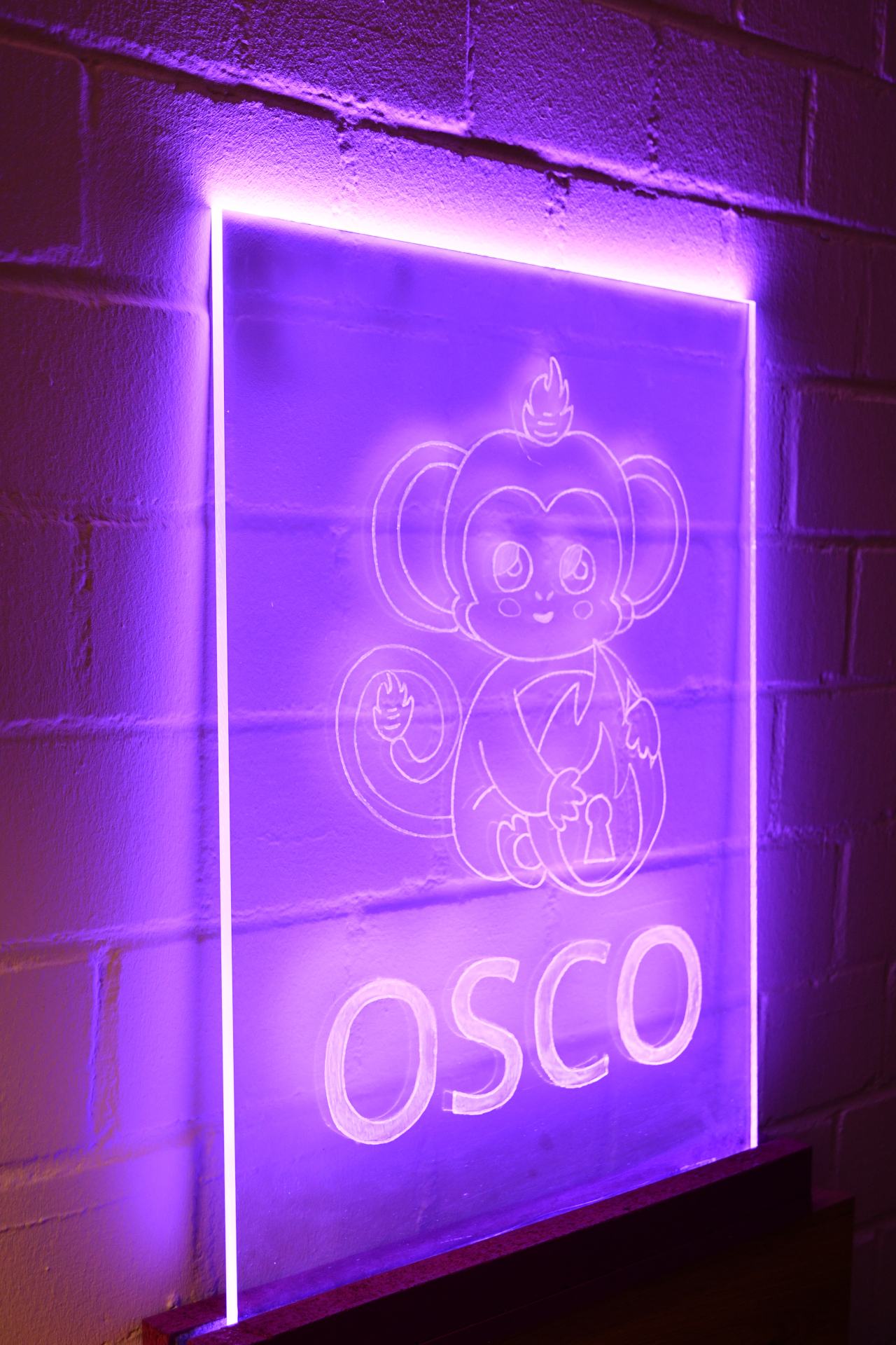 A photo of the osco logo engraved on a big acrylic glas plate shining in bright purple. The rest of the room is dark and the acrylic glas is standin in front of a brick wall generating nice effects with light and shadow.