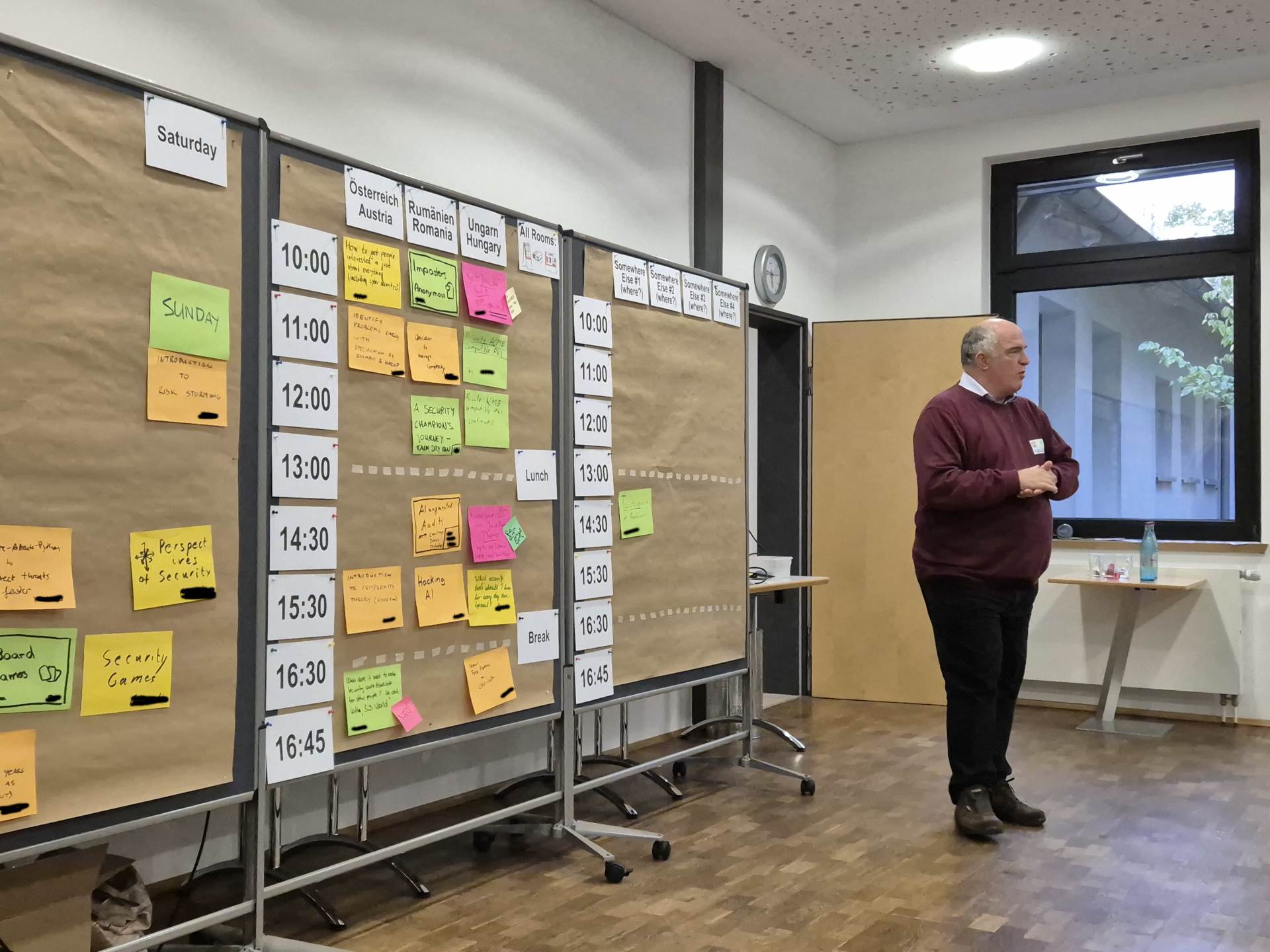 A photo the session planning for the first day of osco 2024. On the left wall are parked slots, that could be happening. On the middle wall are the offered sessions in three parallel rooms. On the right wall is the planning for the next day. One sessions is already there. On the right site of the corner stands a man with red sweater explaining. Your can't see the persons he is talking to. 