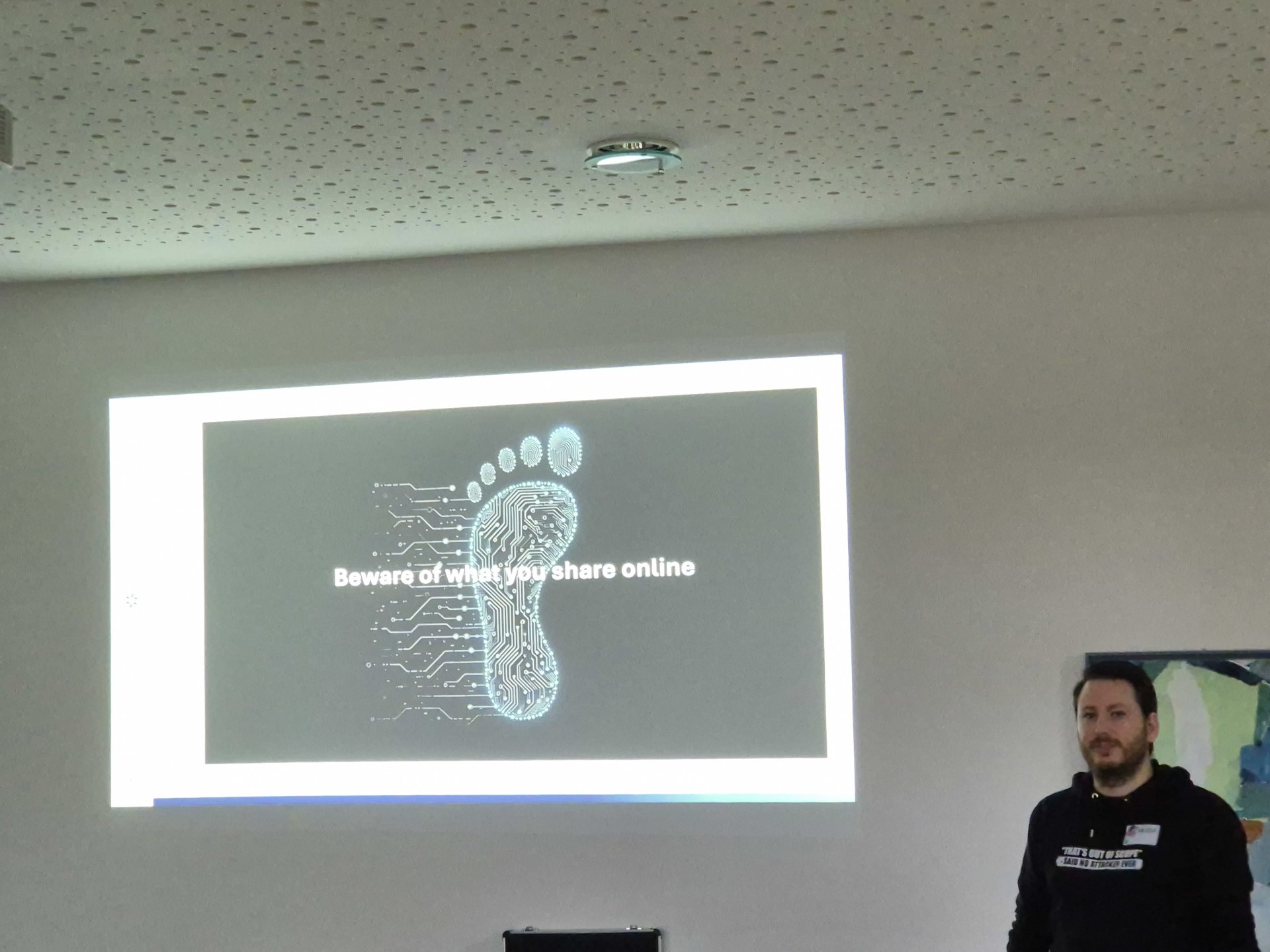 The picture shows part of a presentation. A person in a black hoody with print is in the right part of the picture. Next to hin is the screen with a digital footprint and the text 'beware what you share online'.