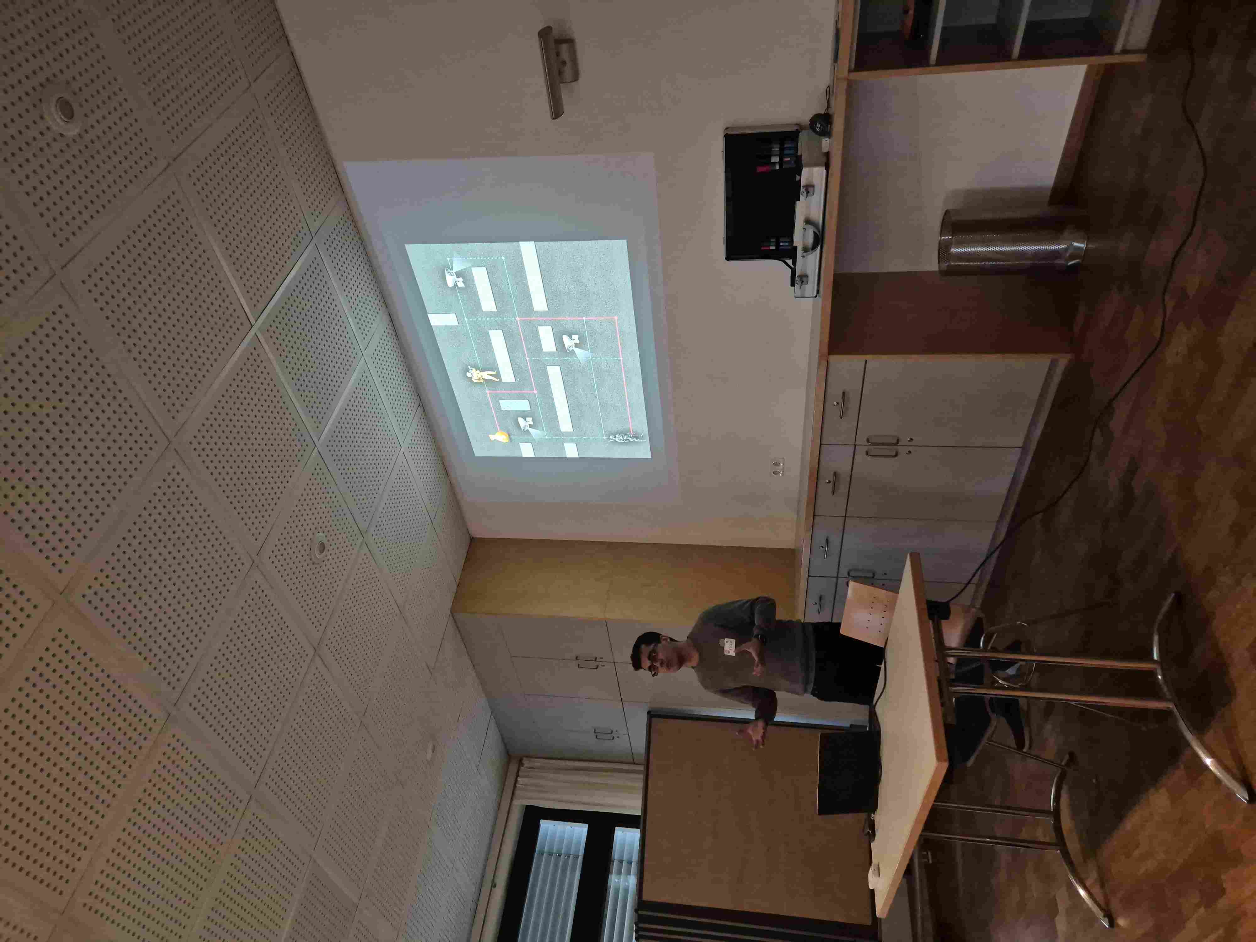 A person with glasses explaining a presentation about deception stands behind desk. In the background is a presentation on the wall with a schema of an intrusion scenario.