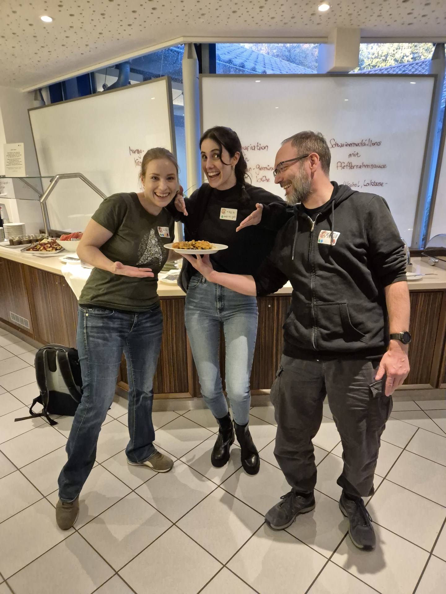 The picture shows three happy people in front of the buffet with great food. They are pointing at a plate.