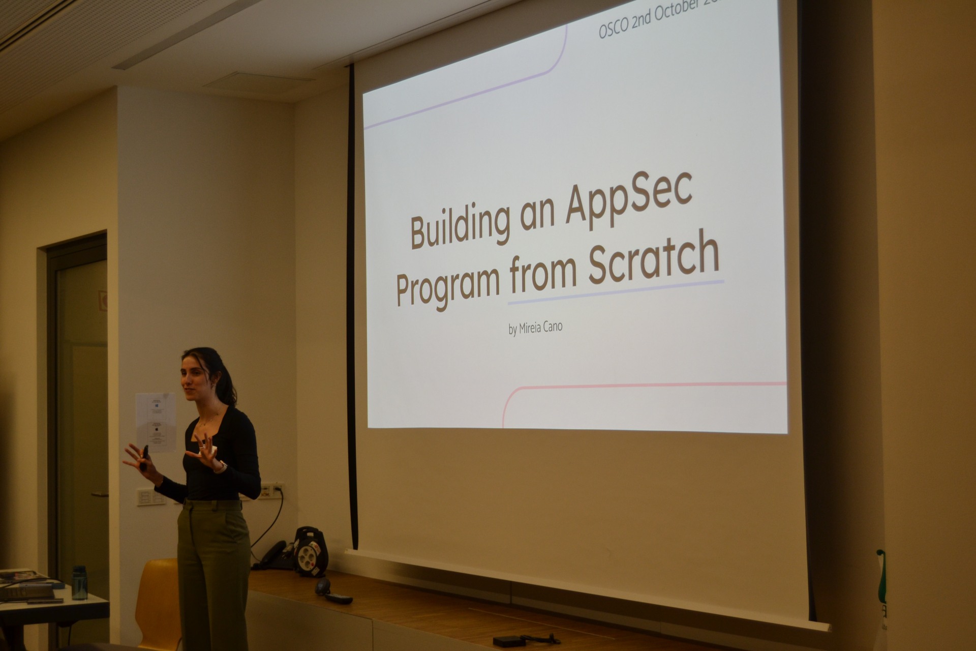 The picture shows aa scene from the keynote 'building appsec program from scratch'. Mireia, the speaker, is talking in the front. In the background is the title page of the presentation on the screen.