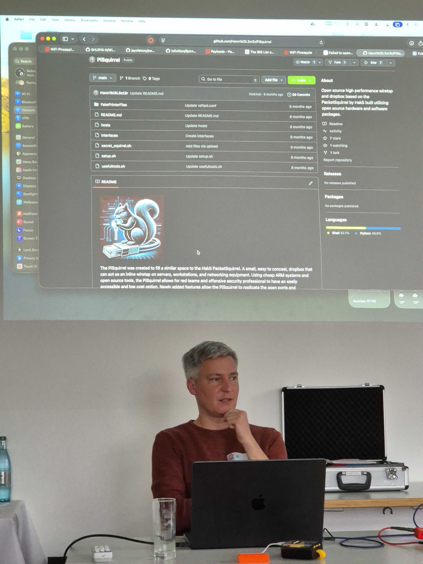 The picture shows part of a presentation. The person behind the laptop has some hacking gadgets at the table. In the background is the screen with some open git repositories.